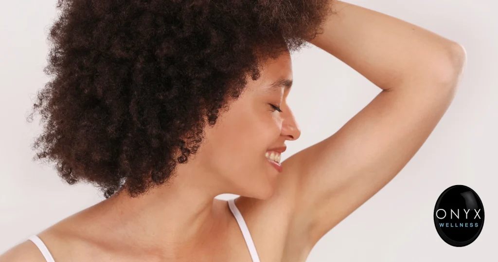 Smiling woman with curly hair raising her arm to reveal a smooth, hair-free underarm, highlighting the results of laser hair removal treatment.